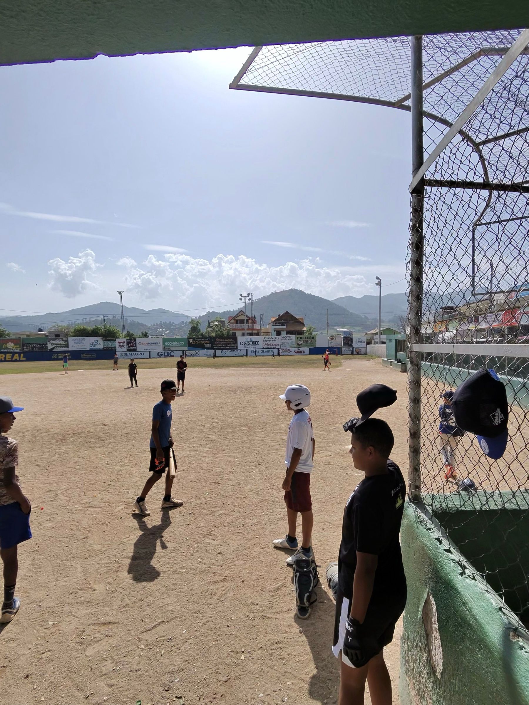 View from the dugout through the fence: kids waiting to hit, caps hanging on the chain-link
